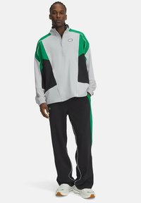 Veste zippée gris clair avec des accents verts et noirs, associée à un pantalon noir avec un passepoil blanc et une coupe décontractée. Des baskets complètent le look.