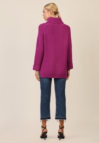 Gruby, oversize sweter w intensywnym purpurowym kolorze, z wysokim kołnierzem i podwiniętymi mankietami, zestawiony z krótkimi dżinsami i butami na obcasie.