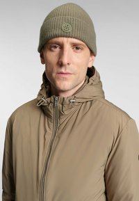 Giacca beige con zip e colletto alto, con regolazione a toggle; berretto a coste verde oliva con una patch circolare del logo. Texture liscia.