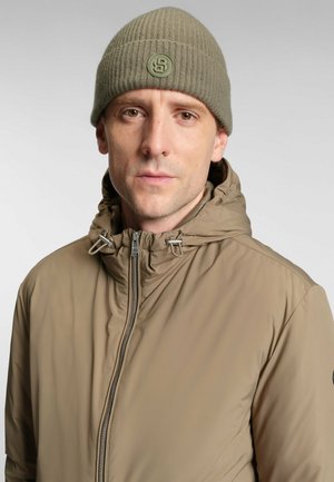 Beige ritssluiting jack met een hoge kraag en toggleregeling; geribde olijfgroene beanie met een cirkelvormig logo patch. Soepele textuur.