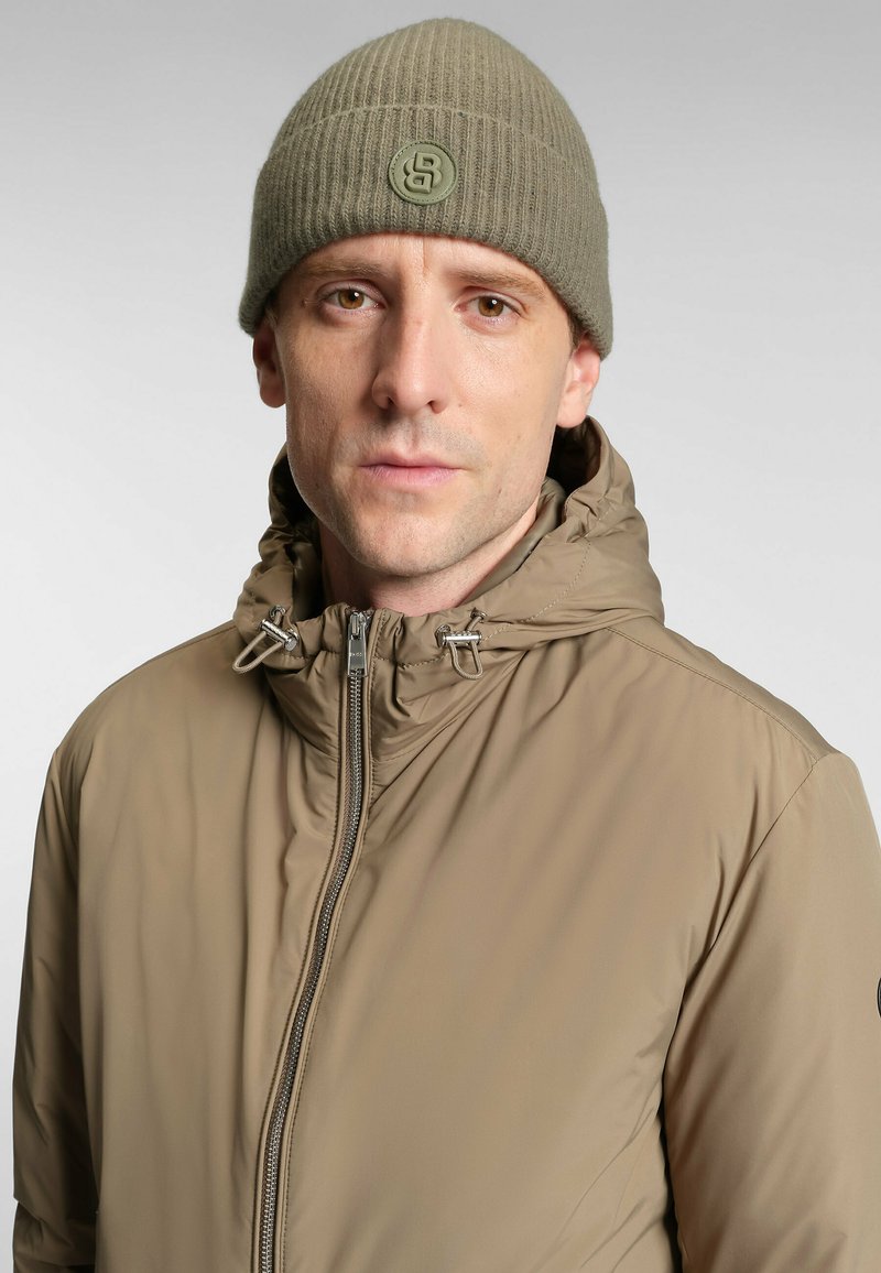 Giacca beige con zip e colletto alto, con regolazione a toggle; berretto a coste verde oliva con una patch circolare del logo. Texture liscia.