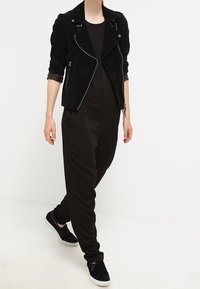 Schwarze Wildleder-Bikerjacke mit Reißverschlusstaschen, kombiniert mit einem lockeren schwarzen Overall und schwarzen Slippern für einen monochromen Look.