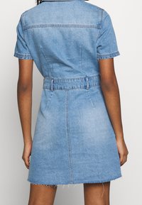 Niebieska sukienka dżinsowa z pasem w talii, krótkimi rękawami, kołnierzykiem i postrzępionym dołem. Posiada dopasowaną sylwetkę i klasyczne przeszycia.