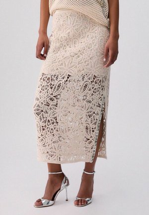 Gonna midi beige in pizzo all'uncinetto con spacco laterale indossata con sandali argentati con tacco alto e cinturino alla caviglia, mostrata su una persona in piedi.