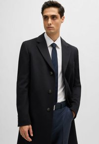 Cappotto in lana nera con taglio dritto, design a un petto, quattro bottoni e revers a punta. Indossato sopra una camicia bianca e una cravatta blu.