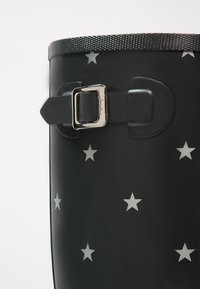 Botas de goma negras con un acabado brillante, decoradas con estrellas blancas y con una hebilla metálica plateada en una correa cerca de la parte superior.