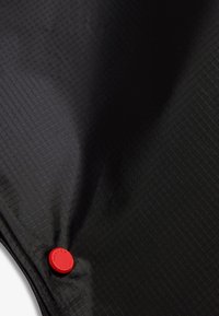 Μαύρο υφασμάτινο ύφασμα με μικρό κόκκινο κυκλικό κουμπί που φέρει ανάγλυφα τα λόγια "Hunter Original" κοντά στην κάτω αριστερή γωνία.