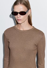 Brauner Langarmoberteil aus einem weichen, strukturierten Stoff. Schwarze, kantige Sonnenbrille mit dunklen Gläsern. Schlichtes Design mit Rundhalsausschnitt.