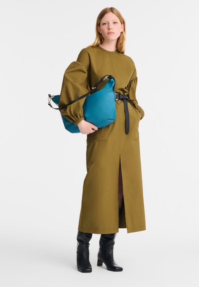 Olivengrünes langes Kleid mit Puffärmeln, seitlichem Schlitz und schwarzem Gürtel; Model hält eine türkisfarbene Tasche und trägt schwarze Absatzstiefel.