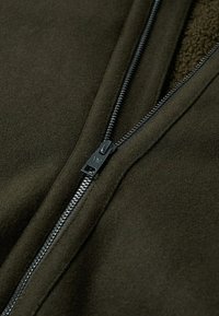 Giacca in fleece verde scuro con chiusura a zip liscia, caratterizzata da una texture morbida e un sottogola di colore contrastante. La cucitura visibile accentua il design.