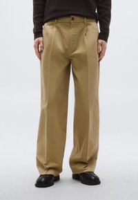 Μπεζ παντελόνι με φαρδιά πόδια και πιέτες μπροστά, με ένα κουμπί και θηλιές για ζώνη, συνδυασμένο με μαύρα δερμάτινα παπούτσια.