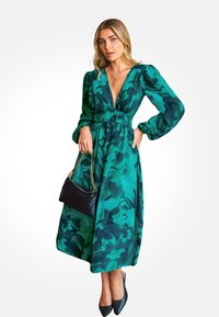 Zielona sukienka w wzory z głębokim dekoltem w kształcie litery V, bufiaste rękawy, marszczeniem w talii i zwiewnym dołem; zestawiona z czarną torebką i szpilkami.
