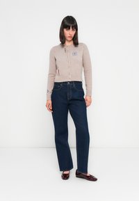 Cárdigan de punto beige con botones, con acentos bordados. Combinado con unos vaqueros de pierna ancha de color azul oscuro y zapatos burdeos brillantes.