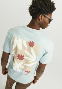 Camiseta de manga corta azul claro con un diseño botánico impreso en beige y rojo en la parte posterior, cuello redondo y un corte relajado.