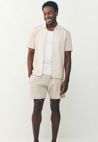 Hombre sonriendo, vistiendo una camisa de cremallera de manga corta beige sobre una camiseta blanca, y pantalones cortos beige a juego, frente a un fondo claro y liso.