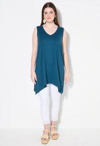 Tunic senza maniche color teal con scollatura a V, design fluido e orlo asimmetrico. Abbinata a pantaloni capri bianchi con bordi in pizzo e sandali espadrilles color tan.