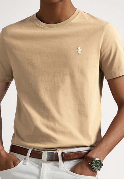 Μπεζ βαμβακερό μπλουζάκι με στρογγυλή λαιμόκοψη και μικρό λευκό λογότυπο στο αριστερό στήθος, συνδυασμένο με καφέ ζώνη και πράσινο ρολόι.