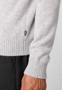 Pull en laine gris clair avec un ourlet et des poignets côtelés. Petite étiquette noire affichant le nom de la marque. Main partiellement visible avec un pantalon sombre.
