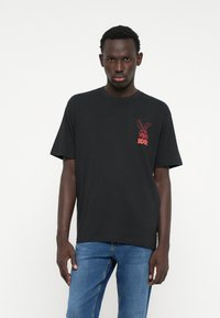 Schwarzes Baumwoll-T-Shirt mit rundem Halsausschnitt und kurzen Ärmeln. Hat ein rotes Motiv im linken Brustbereich. Wird mit blauen Jeans getragen.