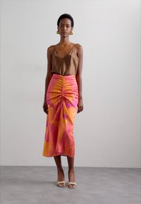 Camisole en satin brun associé à une jupe en soie rose et orange avec des volants et des motifs abstraits. Le modèle porte des talons nus.