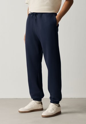 Persona con pantalones deportivos azul marino con puños elásticos y zapatillas blancas de pie frente a un fondo claro y liso.
