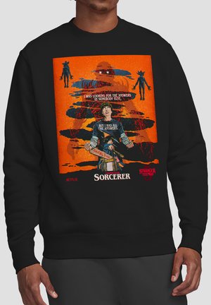 Black sweatshirt with orange graphic from Stranger Things Chapter Four Sorcerer featuring characters, shadowy figures, and text about finding answers.