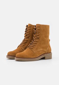 Botas de tornozelo em camurça na tonalidade castanha, com atacadores na parte frontal, bico arredondado e sola plana em borracha. Apresentam costura discreta e uma alça de puxar.