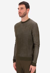 Uomo con un maglione a coste di colore verde oliva e pantaloni abbinati, in piedi di lato davanti a uno sfondo bianco semplice.