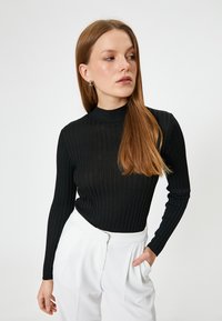 Schwarzes geripptes Langarmoberteil mit hohem Kragen und strukturiertem Stoff, kombiniert mit weißen Hosen. Keine sichtbaren Muster oder Hardware.