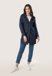 Giacca impermeabile blu navy con cappuccio, dotata di due tasche frontali e chiusura a bottoni, abbinata a jeans blu e sneakers bianche.