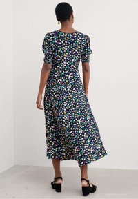 Robe midi florale en tissu foncé avec des motifs multicolores vibrants. Présente des manches bouffantes et une coupe décontractée, associée à des talons noirs épais.