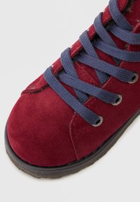 Botim de tornozelo em camurça vermelha com atacadores azul escuro, ilhoses em metal, colarinho almofadado e bico arredondado. Superfície texturizada e sola de borracha.