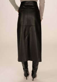 Jupe en cuir noir au toucher lisse, arrivant au genou, dotée d'une taille haute et de détails de couture à l'arrière, associée à des bottes noires à talons hauts.