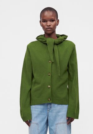 Person iført en grøn knap-karigan med et matchende strikket tørklæde bundet om halsen og lyseblå jeans mod en ensfarvet baggrund.