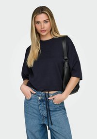 Pull court bleu marine à manches courtes, avec un cordon de serrage à l'ourlet. Associé à un jean en denim taille haute et un sac à bandoulière noir.