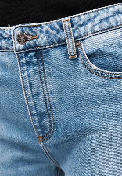 Lyseblå denimjeans i nærbilde som viser midjebånd, knapp med merkenavn, beltehempe og detaljer i sømmen på frontlommen.