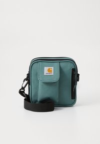 Geantă de umăr verde din material textil, cu un buzunar frontal, curea neagră și închidere cu fermoar. Prezintă un logo pătrat pe partea din față.