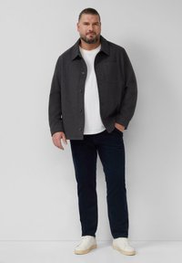 Man met een baard in een donkergrijze knoopjashirts, witte t-shirt, donkere broek en witte sneakers die tegen een effen achtergrond staat.
