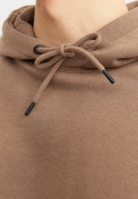 Sudadera marrón con una tela texturizada, que presenta una capucha con cordón y puntas negras. Primer plano de detalles de la tela y el escote.