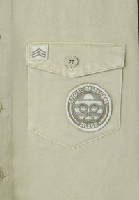 Camisa de algodón beige con bolsillo. Presenta un parche bordado circular con "OPERACIONES ESPECIALES SILBON" y un insignia en forma de chevrón por encima del bolsillo.