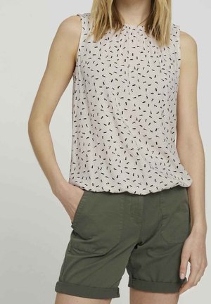 Vrouw draagt een mouwloze witte blouse met een klein zwart patroon en groene opgerolde korte broek, één hand in de zak, tegen een effen achtergrond.