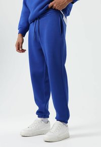 Blue sweatpants made of textured fabric featuring side pockets, elastic cuffs, and a drawstring waistband, paired with white sneakers.