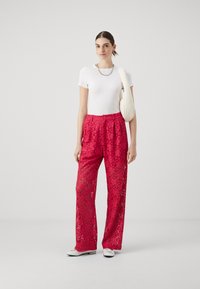 Pantaloni larghi rossi in pizzo con motivi floreali ritagliati, abbinati a una maglietta bianca aderente e a una borsa a tracolla crema. Le scarpe bianche completano il look.