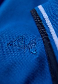 Niebieska tkanina o strukturalnym splocie zdobiona jest haftowanym logo. Czarny zamek błyskawiczny z białymi i niebieskimi paskami akcentuje projekt.