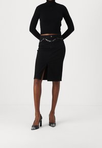 Mujer vestida con un top negro de manga larga y cuello alto, una falda negra hasta la rodilla con abertura frontal y cinturón de cadena plateada, y zapatos de tacón alto negros.