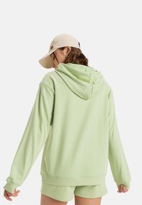 Felpa verde chiaro con cappuccio e maniche lunghe, realizzata in tessuto morbido. Abbinata a pantaloni corti coordinati, lo sfondo è bianco semplice.