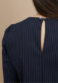 Navyblaues Kleid mit vertikalen weißen Streifen, das an der Rückseite eine Schlüssellochöffnung hat, die mit einem runden schwarzen Knopf befestigt ist, und leicht gepufften Schulternähten.