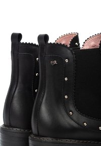 Botines negros con lados de cuero, paneles elásticos y un borde ondulado. Presentan tachuelas metálicas y un pequeño charm en forma de corazón en un lado.