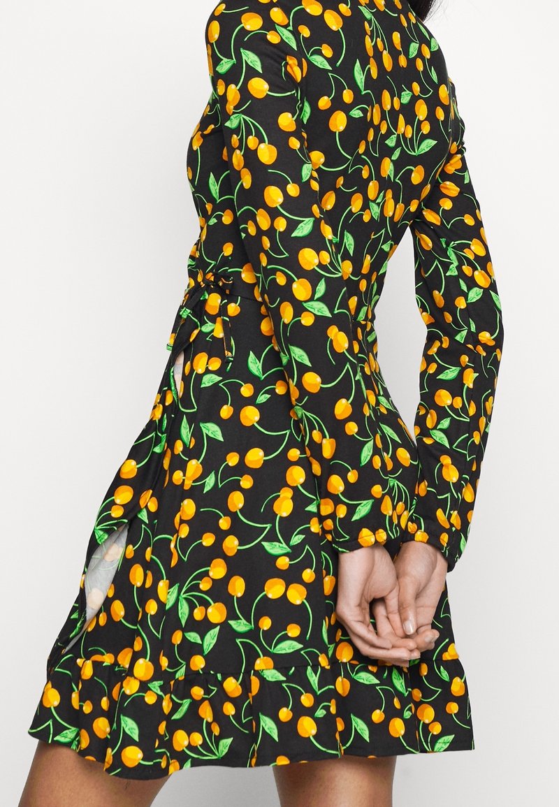 Femme portant une robe noire à manches longues avec un motif de cerises jaunes et de feuilles vertes, mains jointes dans le dos.