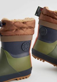Bottes imperméables avec un dessus en nylon matelassé marron, une partie inférieure en caoutchouc vert et marine, une semelle texturée et des symboles météorologiques en relief sur les côtés.
